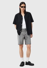 Kortärmad svart skjorta med knappar, vit t-shirt med rund hals, grå denimshorts, svarta skor och solglasögon. Enfärgad bakgrund.