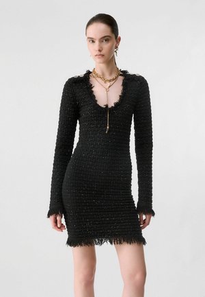 Femme portant une robe mini noire ajustée à manches longues texturée avec des bords frangés et des colliers dorés superposés sur un fond clair uni.