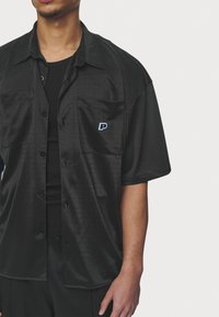 Black short-sleeve shirt made of smooth, glossy fabric, featuring two front pockets and a small blue logo on the left pocket.