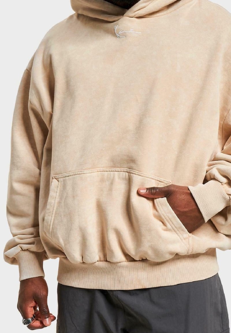 Sudadera con capucha beige hecha de tela suave, con un bolsillo frontal tipo canguro y puños y dobladillo acanalados. Textura sutil y detalle de costuras.