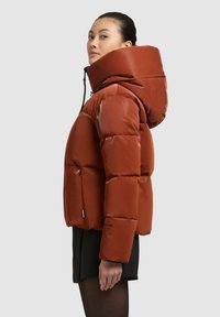 Pūsta oranžinės ir rudos spalvos striukė su aukštu apykakliu ir dideliu gaubtu, kurioje yra užtrauktuku uždaromos priekinės ir šoninės kišenės, blizgus paviršius ir logotipo detalė.