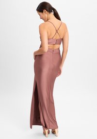 Lang, aansluitend jurkje in stof van stof in een vergrijsde rozenkleur. Heeft kruisende schouderbandjes, een zijsplit en een verborgen ritssluiting aan de achterkant. Zachte textuur.