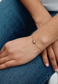 Guldarmbånd med en hjerteformet vedhæng på en fin kæde, båret på en hånd, der hviler på en blå denimoverflade.