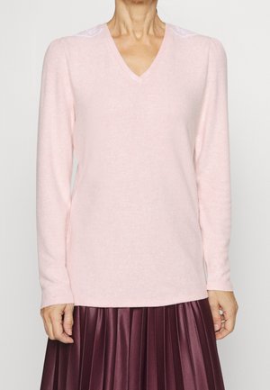 Femme portant un pull rose pâle à col en V avec des détails en dentelle sur les épaules et une jupe plissée violet foncé.