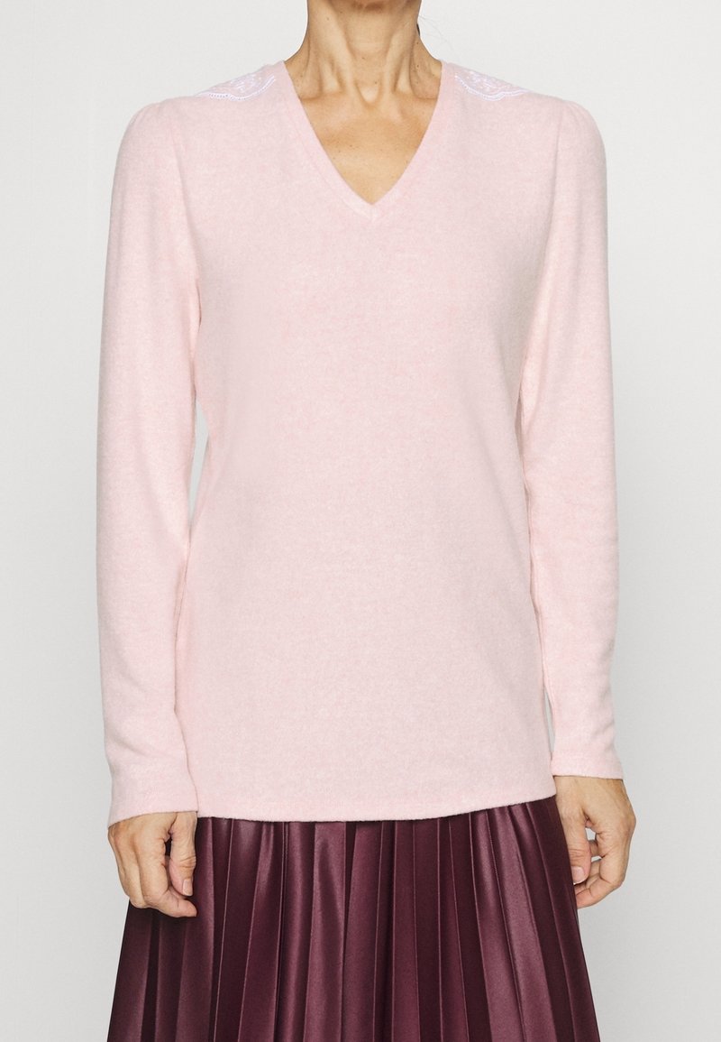 Femme portant un pull rose pâle à col en V avec des détails en dentelle sur les épaules et une jupe plissée violet foncé.