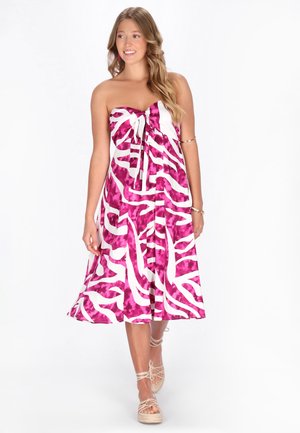 Giovane donna sorridente, che indossa un vestito midi senza spalline rosa e bianco con motivo a fantasia e nodo sul davanti, e sandali beige con zeppa e lacci.