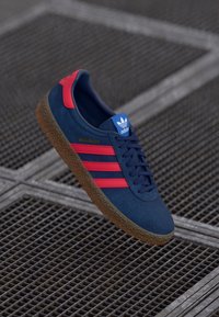 Marineblå ruskinds-sneakers med røde detaljer, tre røde striber på siden, sort gummisål og blåt logo på tungen. Klassisk lav-top design.