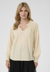 Blouse couleur crème avec des manches longues, décolleté en V et des détails en broderie texturée sur le devant et les poignets. Tissu doux et léger.