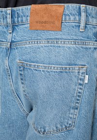 Close-up do bolso traseiro de uns jeans de denim azul-claro com um patch de couro da marca Woodbird e uma etiqueta de tecido branca na borda do bolso.