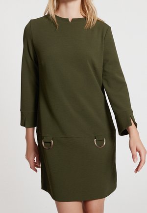 Kvinde iført en olivengrøn langærmet kjole med sølvfarvede D-ring detaljer på skørtet og lette slidsdetaljer ved manchetterne.