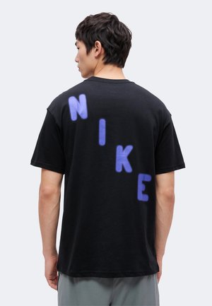Άνδρας που φοράει μαύρο T-shirt με μεγάλα μωβ γράμματα που σχηματίζουν τη λέξη «NIKE» διαγώνια στην πλάτη, στέκεται μπροστά σε ένα απλό ανοιχτόφωτο φόντο.