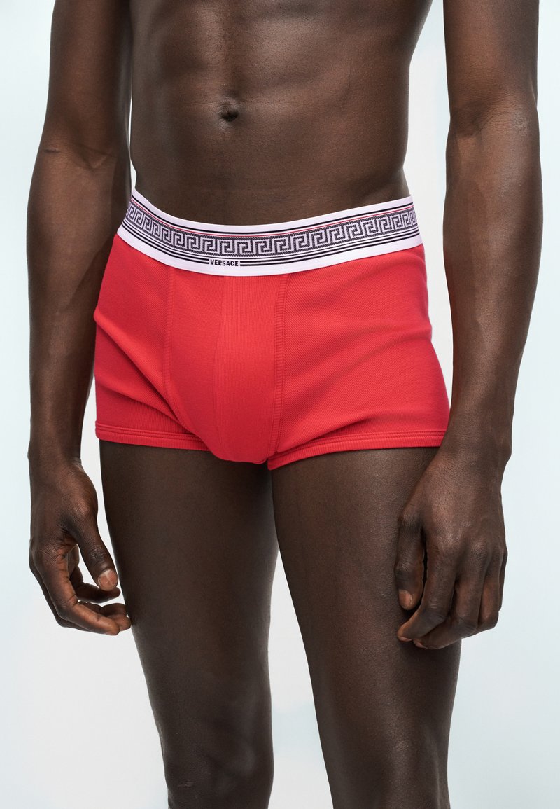 Mannelijk model draagt rode Versace boxershorts met een witte tailleband met een zwart Grieks sleutelmotief.