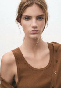 Camisa sin mangas marrón con un escote redondo y un detalle de botones en un lado, hecha de una tela suave, usada por una persona de piel clara y cabello castaño.