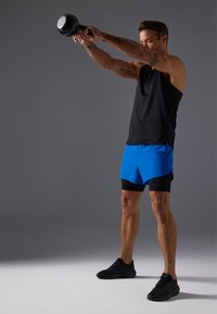 Melna bezgarša, zila šorti, kas uzvilkti virs melnajām kompresijas šortiem. Melni sporta apavi. Tur rokās kettlebellu ar paceltām rokām.