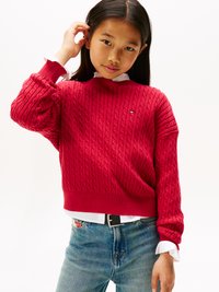 Rød kabelstrikket genser med ribbestrikkede mansjetter og kant, brukt over en hvit skjorte med krage. Lyseblå jeans og et svart belte med sølvspenne.
