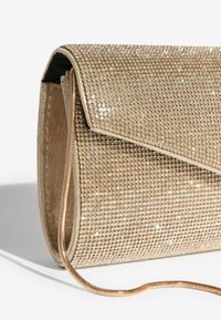 Geantă tip clutch de culoare aurie, acoperită cu strasuri sclipitoare, având un capac care se pliază peste și o curea metalică în lanț. Suprafeța texturată reflectă lumina.