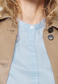 Beige läderjacka med slag, tillsammans med en ljusblå skjorta med knappar och rund krage, som har två vita knappar vid halsen.