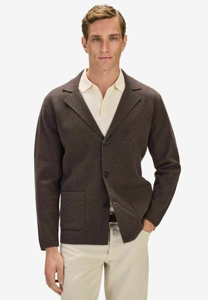 Man i en mörkbrun avslappnad kavaj över en cremefärgad pikétröja med beige byxor, stående med ena handen i fickan.