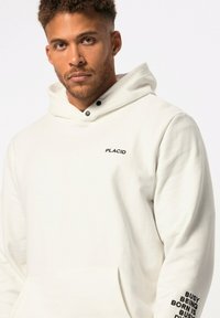Sudadera blanca con capucha ajustable, texto negro "PLACID" en el pecho y "BUSY BEING BORN IS BUSY" a lo largo de la manga. Textura de tela suave.