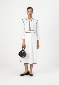 Robe midi blanche avec des bordures noires, col à boutons, manches longues et taille cintrée. Le mannequin tient un petit sac à main noir et porte des ballerines noires.