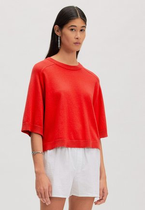 Vrouw met lang donker haar draagt een rode losse trui met korte mouwen, witte korte broek, zilveren oorbellen en een armband, staand tegen een effen achtergrond.