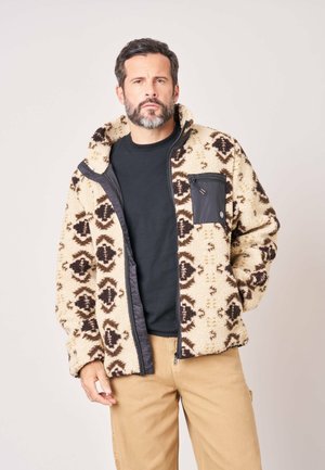 Man draagt een beige fleecejack met patroon over een zwart shirt en beige broek, staand tegen een effen lichte achtergrond.
