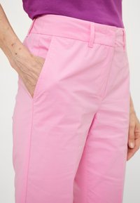 Persoon in lichtroze broek en een paarse top, met één hand gedeeltelijk in de voorzak van de broek.