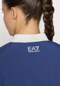 Navy blue jacket featuring a white collar and EA7 Emporio Armani logo on the back. Smooth texture and lightweight material.