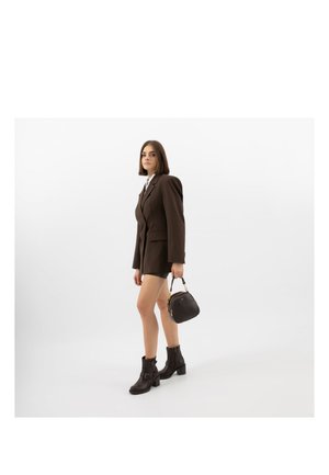 Woman in brown blazer, black ankle boots, carrying a black handbag, standing against a plain white background.