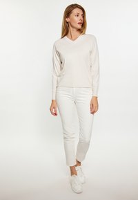 Vaaleanbeige, V-pääntiepusero, jossa on pitkät hihat, yhdistetty valkoisiin slim-fit-housuihin ja valkoisiin tenniskenkiin. Sileä pinta, rento muotoilu.