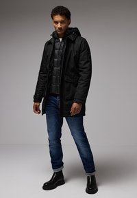 Černá voděodolná parka s kapucí, vrstvená nad černou bundou s péřovou výplní. Kombinována s modrými džíny a černými kotníkovými botami, postavená proti neutrálnímu pozadí.