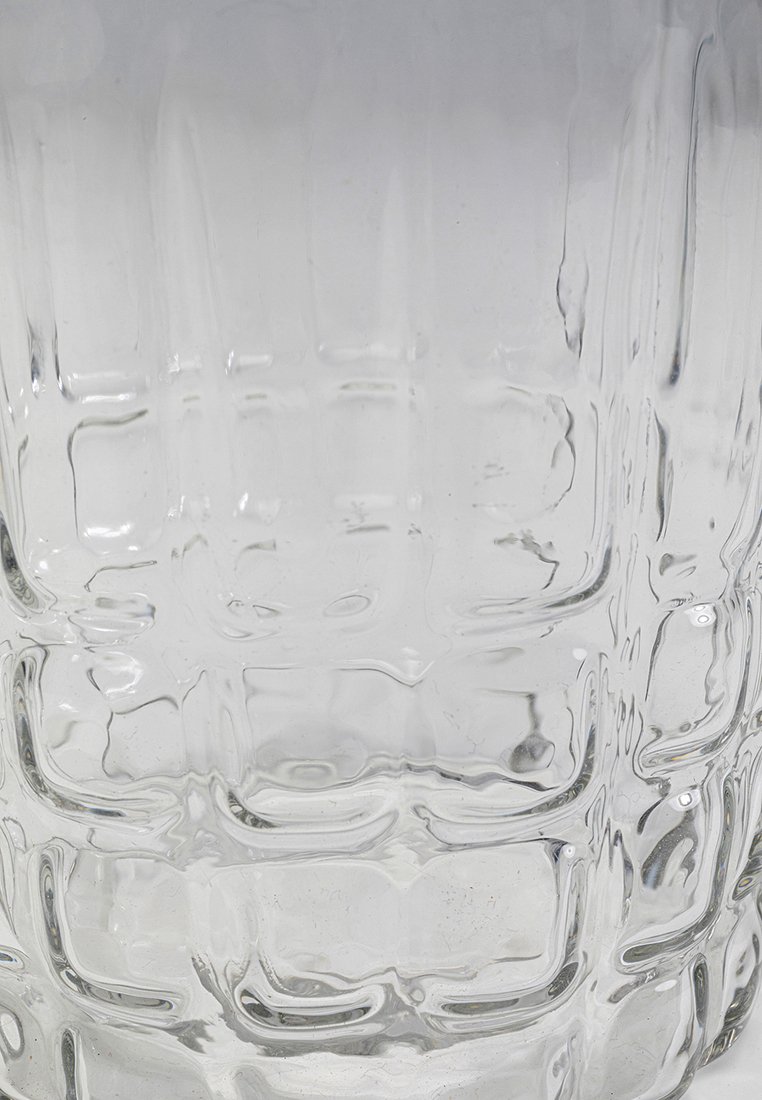 Taza de vidrio transparente con una superficie texturizada que presenta líneas horizontales y verticales, forma cilíndrica y borde suave.