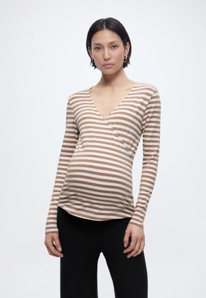 Femeie însărcinată cu păr scurt și închis la culoare, purtând un top cu mâneci lungi în dungi bej și albe și pantaloni negri, stând în picioare pe un fundal simplu.