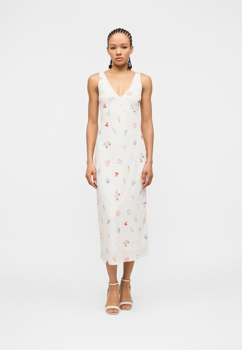 Γυναίκα που φοράει ένα λευκό midi φόρεμα χωρίς μανίκια με πολύχρωμα μικρά λουλουδένια και θαλασσινά μοτίβα, στέκεται με ουδέτερη έκφραση και άσπρα τακούνια.