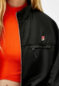 Närbild av en person som bär en ljusorange ribbad crop top under en svart jacka med dragkedja och en liten röd och vit logotyp på bröstet.