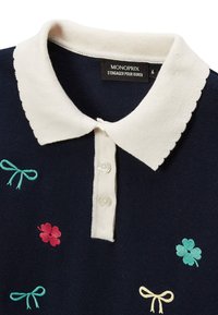 Polo bleu marine avec un col festonné blanc, une patte de boutonnage à trois boutons, et des broderies de nœuds et de fleurs en vert, rose et jaune.