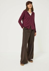 Cardigan bordeaux à manches longues avec des boutons, associé à un pantalon large marron. La tenue comprend une casquette et des chaussures décontractées à semelle épaisse.