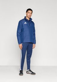 Μπλε μπουφάν με μόνωση και φερμουάρ, συνδυασμένο με αντίστοιχα παντελόνια φόρμας με άσπρες ρίγες στο πλάι, και μαύρα αθλητικά παπούτσια με άσπρες λεπτομέρειες.