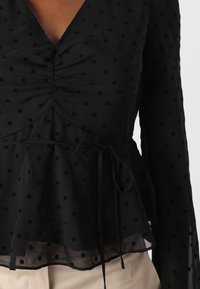 Blusa nera trasparente a maniche lunghe con pois, con fronte arricciato, vita annodabile a portafoglio e orlo a strati, abbinata a pantaloni beige.