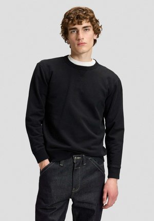 Giovane uomo con capelli ricci che indossa una felpa nera sopra una camicia bianca e jeans scuri, in piedi con una mano in tasca contro uno sfondo semplice.