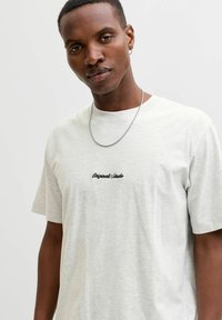 Heled hallidest puuvillane T-särk lühikeste varrukatega, mille keskel on musta tikandiga "Originals Studio" tekst. Lisandiks kettkaelakee.