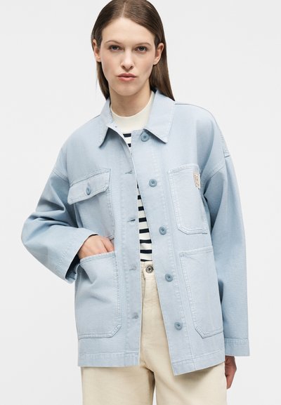 Γυναίκα που φοράει ανοιχτό μπλε τζιν μπουφάν oversized πάνω από ριγέ πουκάμισο και μπεζ παντελόνι, με το ένα χέρι στην τσέπη του μπουφάν, μπροστά σε λευκό φόντο.