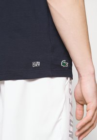 Marinblå bomullströja med "ULTRA DRY" tryck nära fållen och en grön broderad krokodil-logotyp på sidan, slät textur.