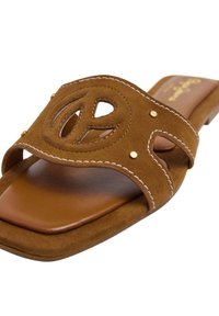 Bruine suede slipper met een uitgesneden vredessymbool, decoratieve stiksels en kleine metalen accenten op de banden. Plat zoolontwerp.