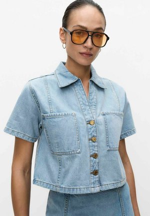 Kobieta nosząca duże okulary przeciwsłoneczne z pomarańczowym filtrem oraz jasnoniebieską kurtkę jeansową z krótkim rękawem, z przednimi kieszeniami i złotymi guzikami.