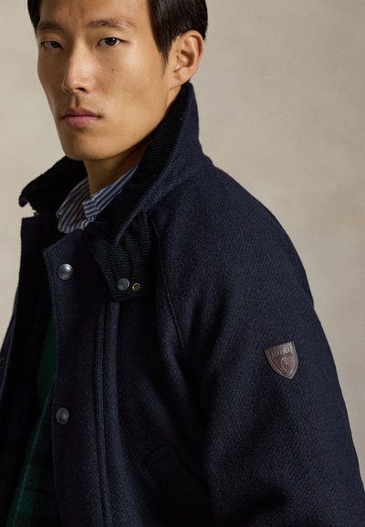 Mörk marinblå ullblandad kappa med knäppning fram, manchesterribb i kragen och lädermärke med logotyp på axeln. Texturerat tyg och skräddarsydd passform.