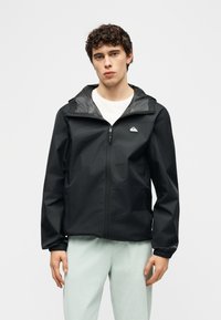 Νέος άνδρας που φορεί μαύρο φούτερ με φερμουάρ και κουκούλα πάνω από άσπρο πουκάμισο και ανοιχτόχρωμο παντελόνι, στέκεται μπροστά σε απλό φόντο.