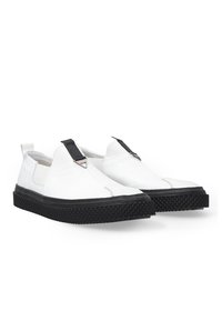 Weiße Leder-Slipschuhe mit einer schwarzen strukturierten Sohle, ausgestattet mit einem schwarzen elastischen Riemen und dreieckigen silbernen Beschlägen im oberen Bereich.