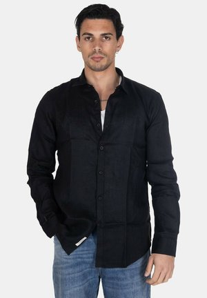 Uomo con capelli corti e scuri e orecchino che indossa una camicia nera con bottoni sopra una maglietta bianca e jeans blu, in piedi davanti a uno sfondo uniforme.