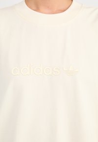 Kremfarget bomulls t-skjorte med en hevet, tonet "adidas" logo og trefoil grafikk i midten. Enkel, glatt tekstur.